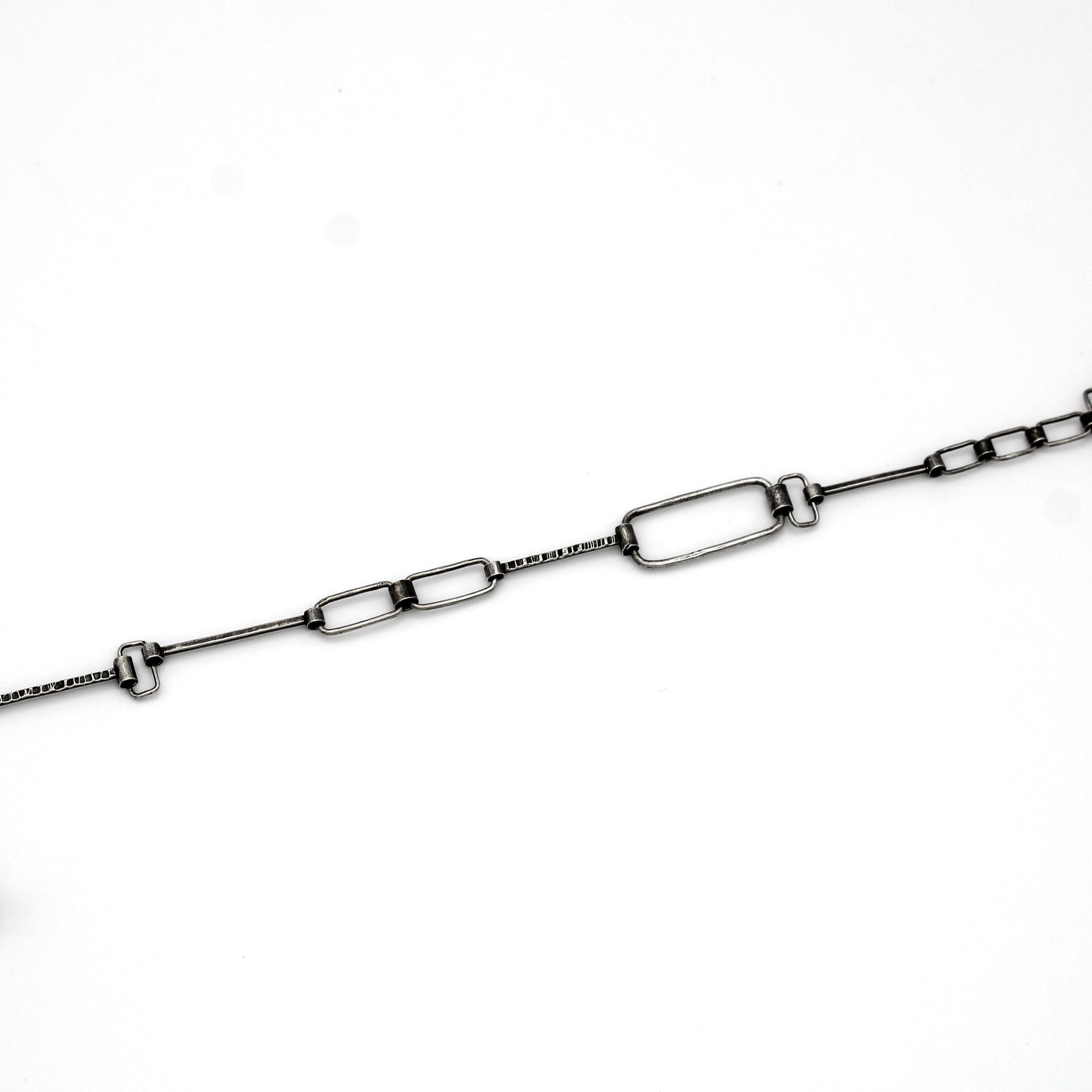 The image shows a close-up of a silver chain with various rectangular and geometric-shaped links. The chain has an industrial and minimalist design, with some links being elongated rectangles, while others are smaller and more compact. The background is white, providing a clean and clear view of the chain's structure and details.