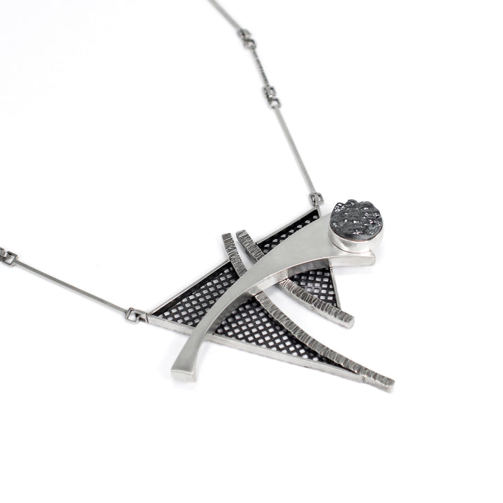  Modernist-inspired sterling silver pendant fashioned in a downward-facing triangle. The triangle is backed by a black mesh and vertically bisected with a widening swoop. A hollow form, mirroring the curve in the triangle, rests horizontally across the pendant, resembling a comet. The broad end of the swoop is on the right, where a 15cm botryoidal hematite is set in fine silver