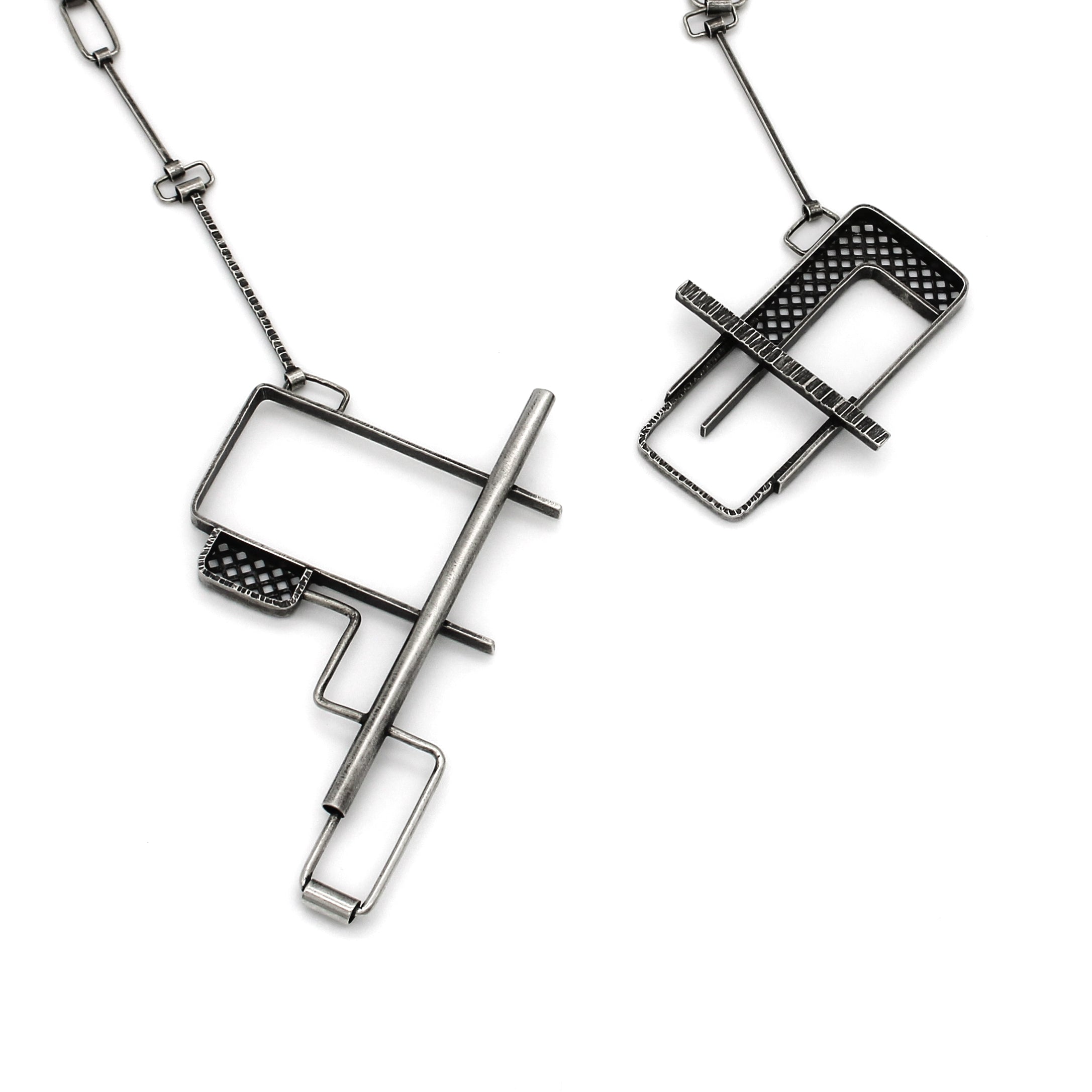 
The image shows two segments of an industrial-style silver necklace with intricate geometric designs. The pendant sections are composed of rectangular and linear shapes, with one piece featuring a hollow, grid-patterned design and the other incorporating a bar-like element. Both pieces are connected by a chain with rectangular links. The image showcases a puzzle clasp mechanism, where the two segments interlock to form a closure. The necklace has a modern, minimalist aesthetic, with attention to bot