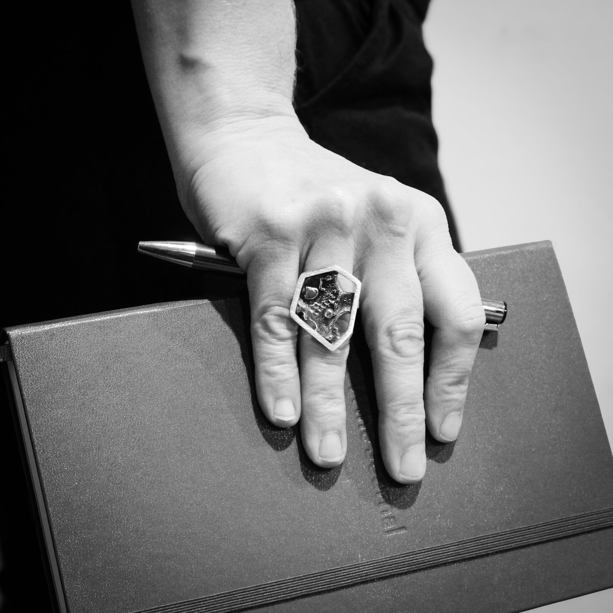 Black and white close-up of a hand holding a book and pen, with a focus on the sterling silver brutalist hexagon ring worn on the ring finger.