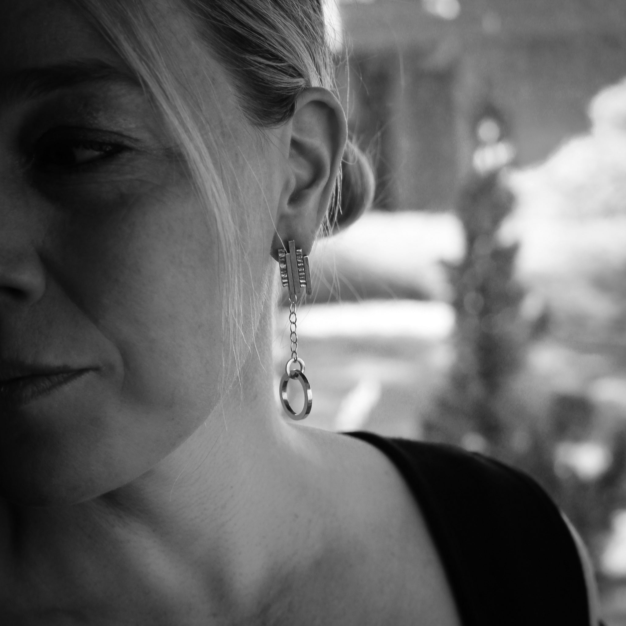 Black and white close-up of a person wearing sterling silver earrings. The earrings feature textured bars at irregular heights, a chain, and a smooth circular hoop. The minimalist design contrasts with the intricate textures, offering a modern yet timeless aesthetic