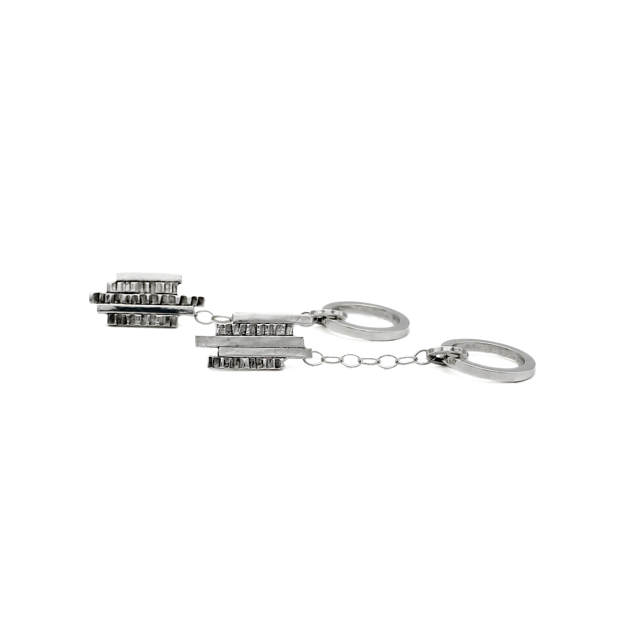 Sterling silver earrings laying on their side, featuring an element of textured bars lined up at irregular heights at the top of the earring, with a delicate chain leading to a smooth circular hoop. The geometric design combines rugged and polished elements, creating a modern and artistic look