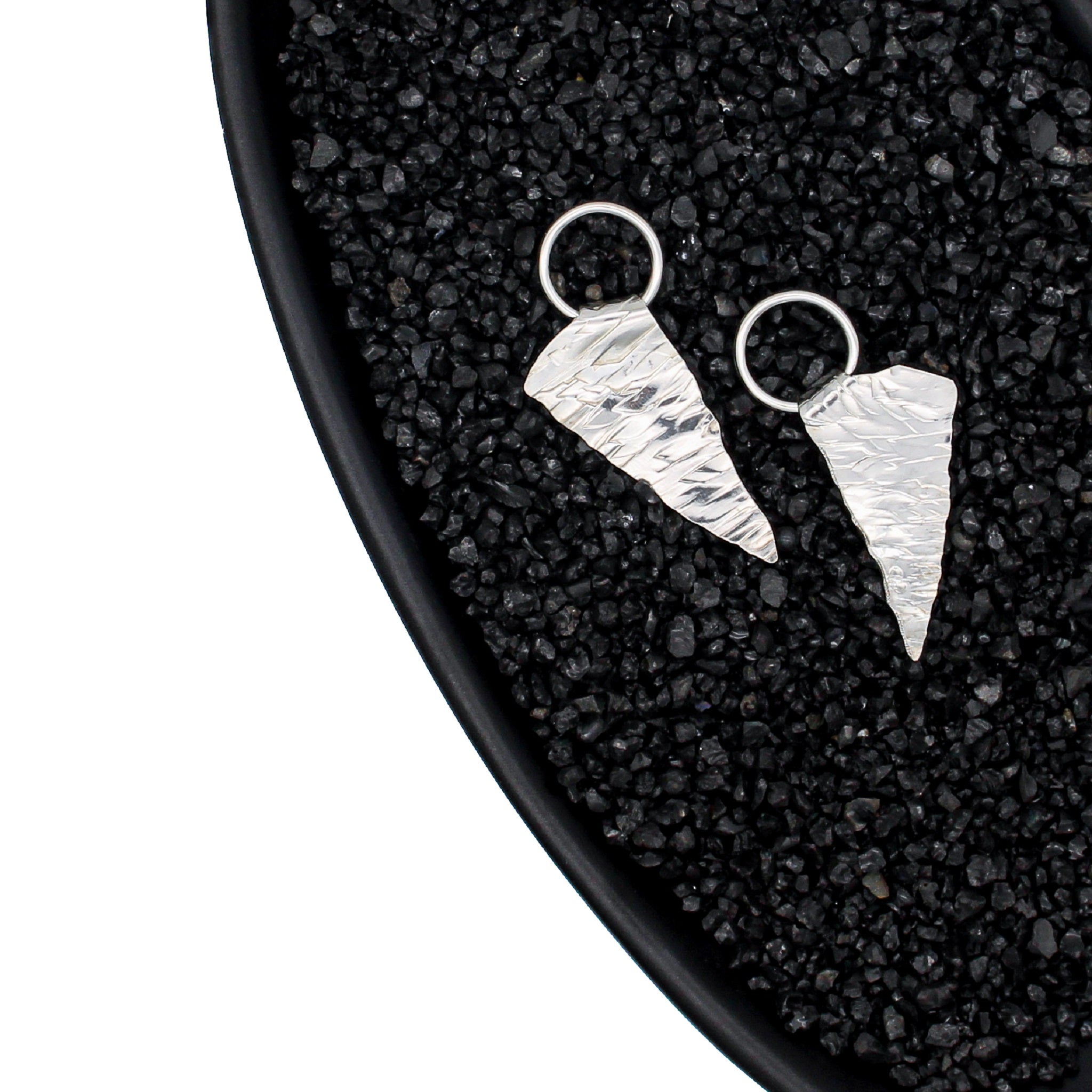 A one of a kind pair of hand fabricated silver earrings, each a rough triangular shape with a textured surface resembling ice. They are suspended from a sterling ring attached to ear posts. The earrings are seen in a black dish, with black pebbles