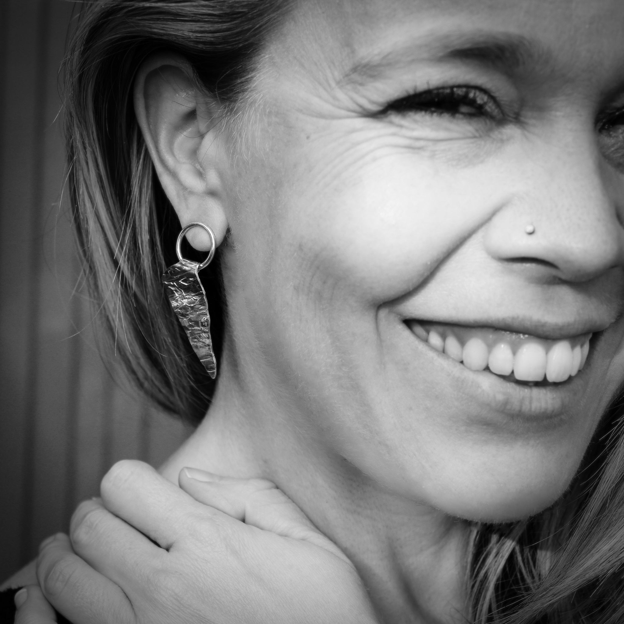 A smiling woman's face, looking at the camera in black and white, wearing a triangular sterling silver earring textured to resemble ice. The earring is suspended from a ring, attached to an ear post."