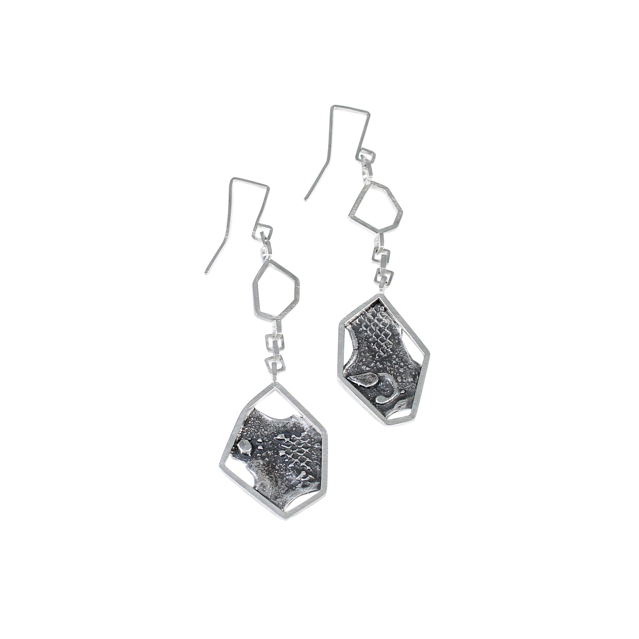 Sterling silver modern dangle earrings, on white background. Each earring has a larger hexagon framing a blackened and brutalist textured backplate. This is attached to some square chain links, and smaller hexagon and finished with a square shaped ear wire.