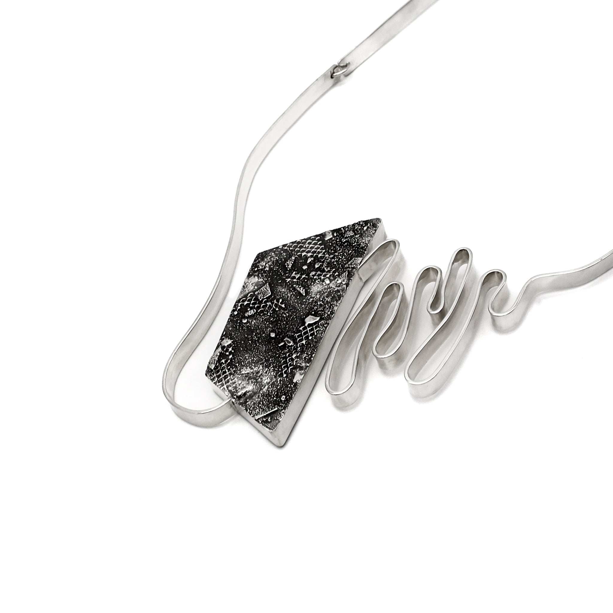 A modernist inspired handmade sterling silver necklace showcasing an undulating bar on the left side, mimicking soundwaves, culminating at the left side of an asymmetric pentagon hollowform. The hollowform has a blackened, heavily textured surface, evoking a brutalist sensibility. Emerging from the bottom of the hollowform, the silver bar creates the illusion of continuity as it curves upward, connecting to a bar and link chain. Pendant is angled and focus is on the textured black hollowform