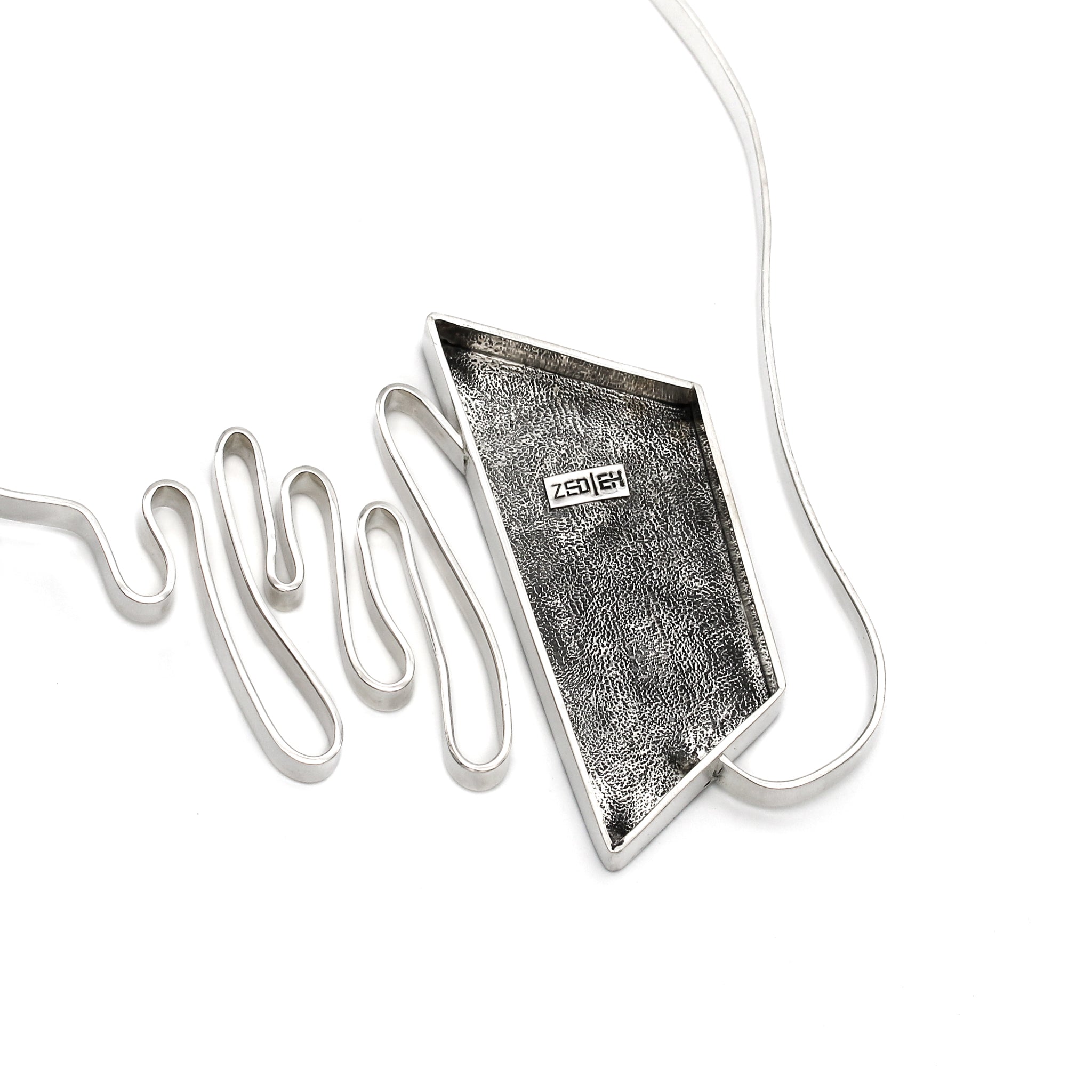 The back of a modernist inspired handmade sterling silver necklace showcasing an undulating bar on the left side, mimicking soundwaves, culminating at the left side of an asymmetric pentagon hollowform. The open back of the hollowform has a blackened stipple texture and the Zed Eh Designs logo can be seen in the center. Emerging from the bottom of the hollowform, the silver bar creates the illusion of continuity as it curves upward, connecting to a bar and link chain. 