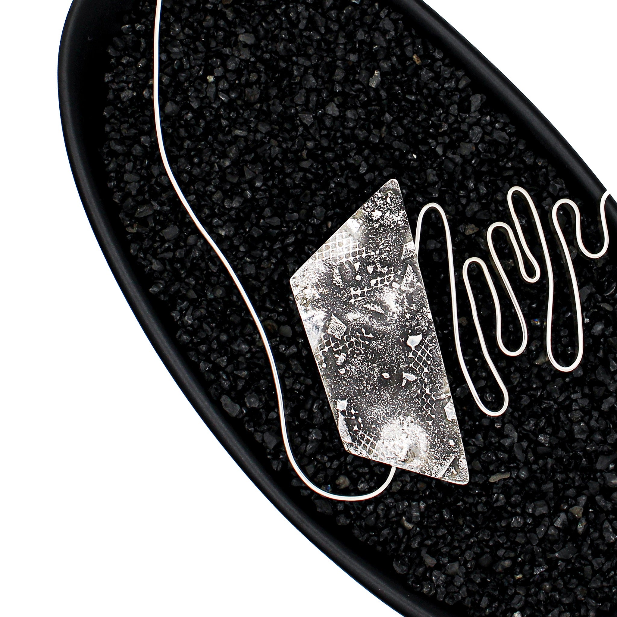 A modernist inspired handmade sterling silver necklace showcasing an undulating bar on the left side, mimicking soundwaves, culminating at the left side of an asymmetric pentagon hollowform. The hollowform has a blackened, heavily textured surface, evoking a brutalist sensibility. Emerging from the bottom of the hollowform, the silver bar creates the illusion of continuity as it curves upward, connecting to a bar and link chain. Pendant is in a black dish with black pebbles. 