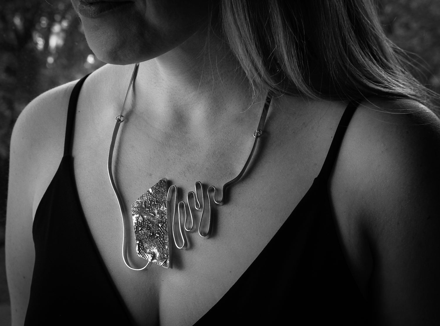 Black and white close-up of woman’s torso, wearing a black, sleeveless shirt. She is wearing a large modern statement pendant, featuring sleek undulating silver bars and brutalist textured hollowform.