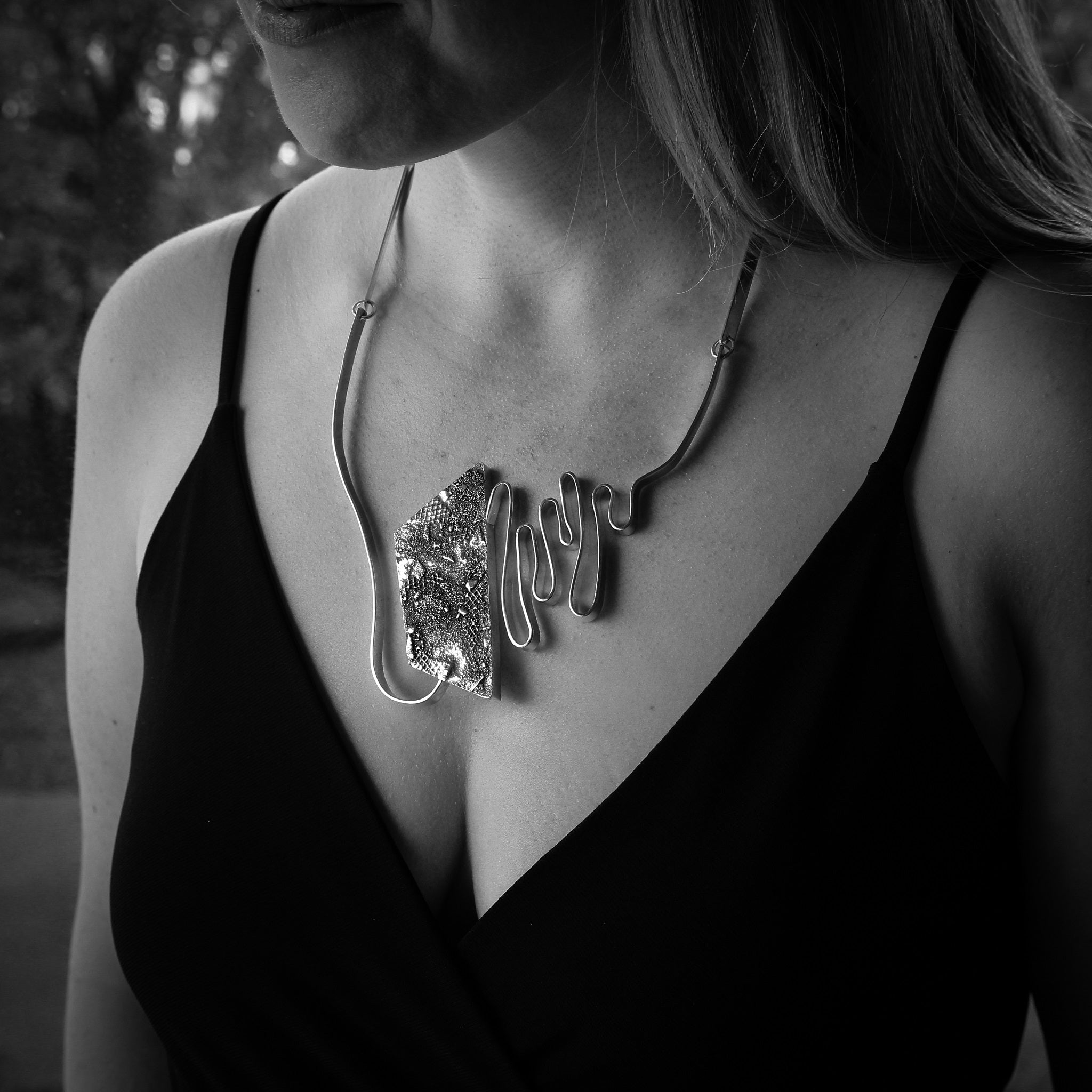 Black and white close-up of woman’s torso, wearing a black, sleeveless shirt. She is wearing a large modern statement pendant, featuring sleek undulating silver bars and brutalist textured hollowform. 