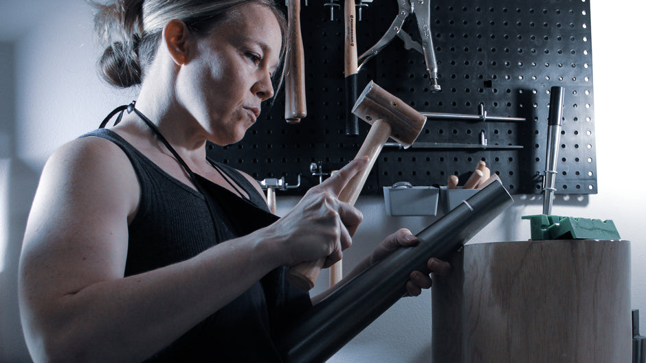 Side profile of woman, using a rawhide mallet, to hammer a curved piece of sterling silver on a large steel bracelet mandrel. The mandrel is propped on a round wooden stump. The woman's hair is tied in a bun, she is wearing safety glasses, and a blue tank top, with a black apron. Various hammers hung on the wall can be seen in the background.