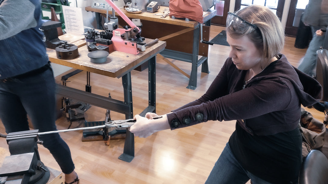 A woman with short dirty blonde hair, wearing blue jeans, a black shirt, and a maroon half-length sweatshirt, is forcefully pulling and twisting very thick sterling silver wire. The wire is anchored to a vice on a wooden table, and she is sing both arms and her full body strength for the task. Her safety glasses are pushed up, securing her hair back. In the background, a bustling silversmithing studio is visible