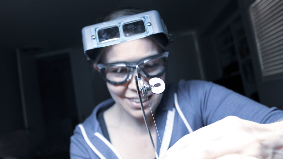 A close-up view captures a woman holding a black Green Lion saw, with the focus centered on the tool. The woman is slightly blurred in the background, wearing safety glasses, with her hair tied back. She has on a blue sweatshirt and displays a cheerful smile