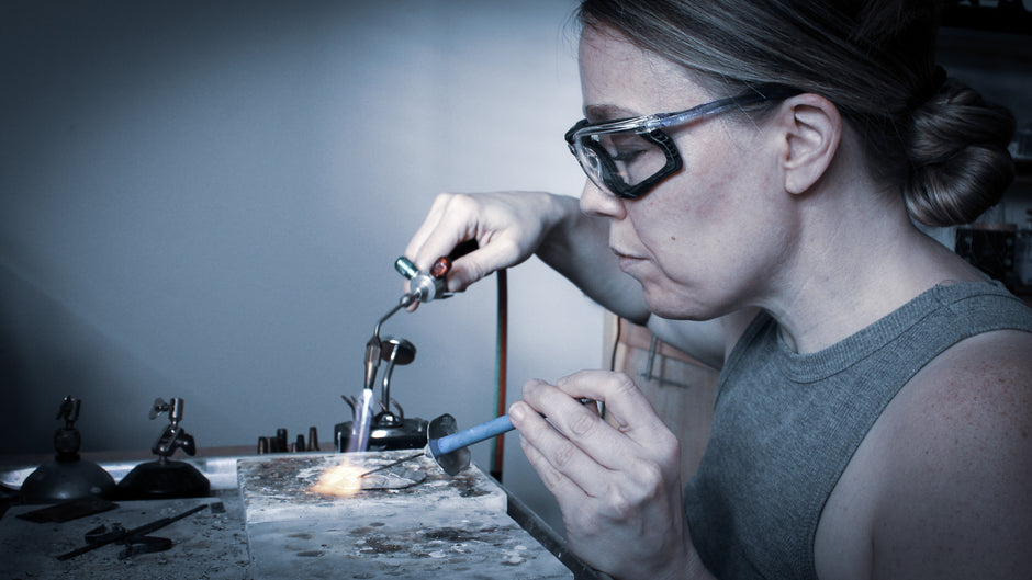 Side profile of a woman engaged in metalworking, holding a lit soldering torch in her right hand and a blue soldering pick in her left. The torch is directed downward toward an oval piece of silver resting on a white solderite board. The pick is maneuvered within the flame. The woman, with her hair tied up, wears safety glasses and a grey tank top. In the background, her workbench is intentionally blurred, emphasizing the focused action at the forefront