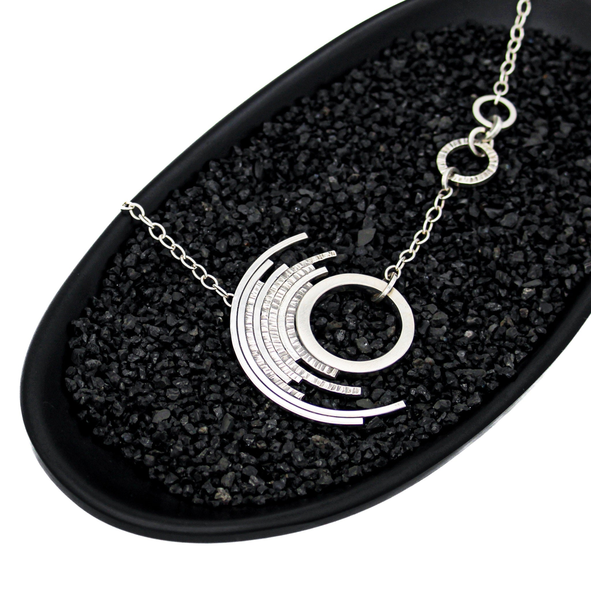 Modern silver asymmetric pendant featuring a medium circle surrounded by 7 concentric circles in a black dish with black stones 