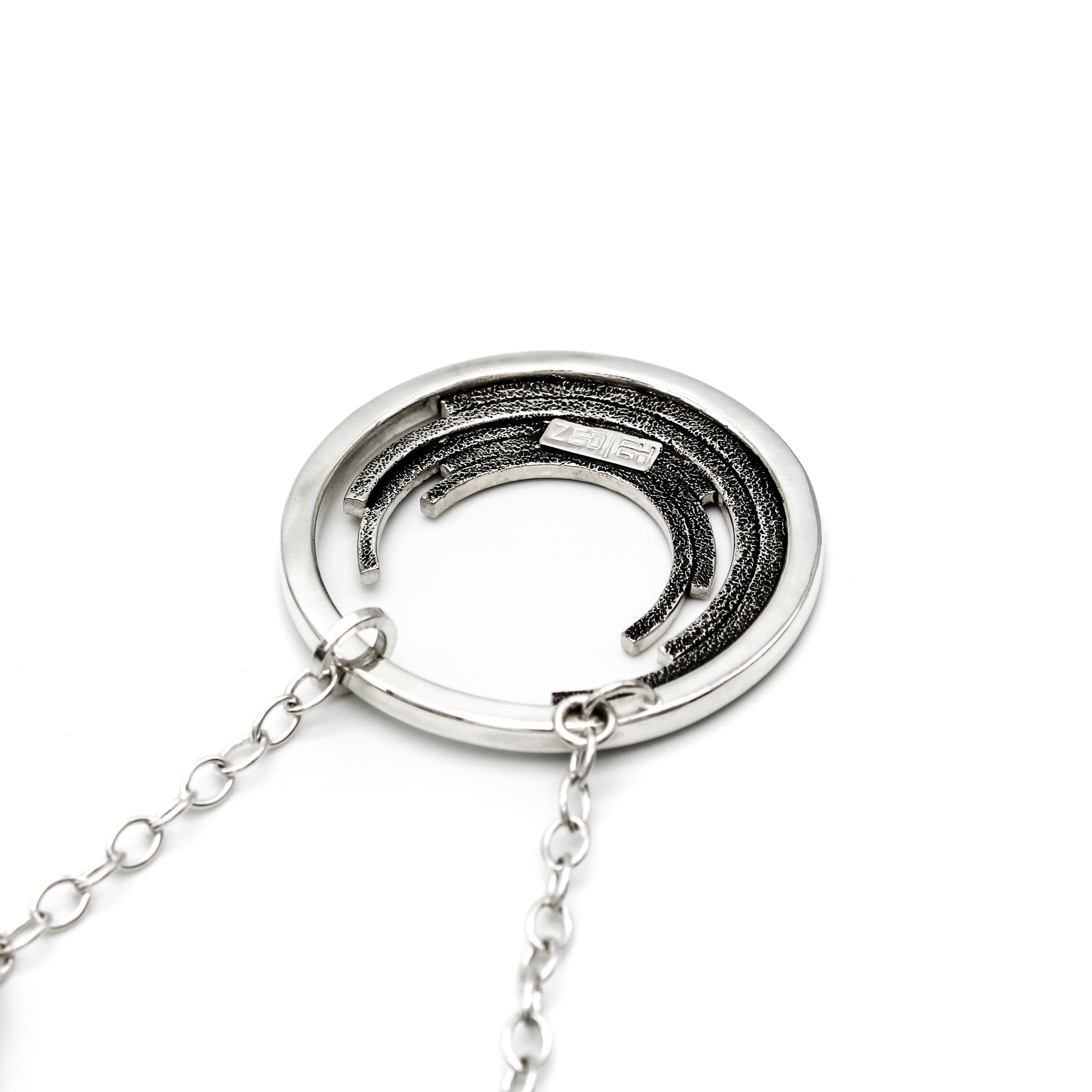 Reverse side of a sterling silver pendant showcasing a sizable circle with five staggered, progressively smaller inner circles. The back of the pendant displays a textured stipple pattern, accentuated by a blackened finish. The Zed Eh Design logo is attached to the center of the pendant's back