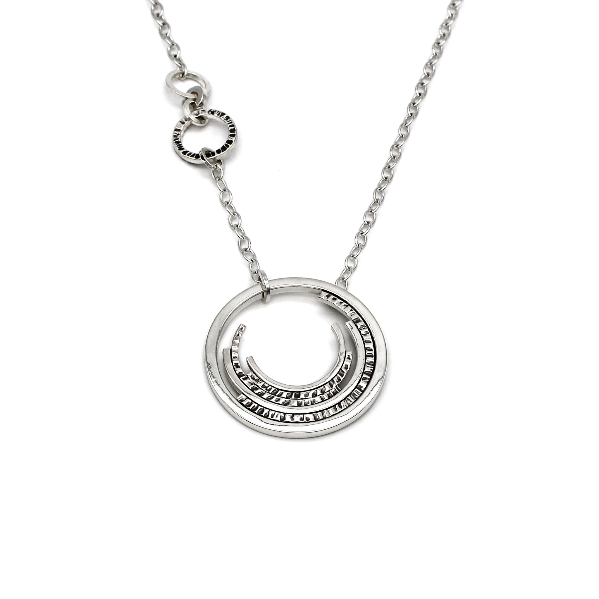 Contemporary sterling silver necklace with a prominent outer circle surrounded by five descending, staggered inner circles. The inner circles showcase varying textures, some chisel-textured and blackened for enhanced visual contrast. Suspended from a sterling silver cable chain, the pendant is complemented by four irregularly sized and textured rings positioned 1.5 inches up the chain on the right side 