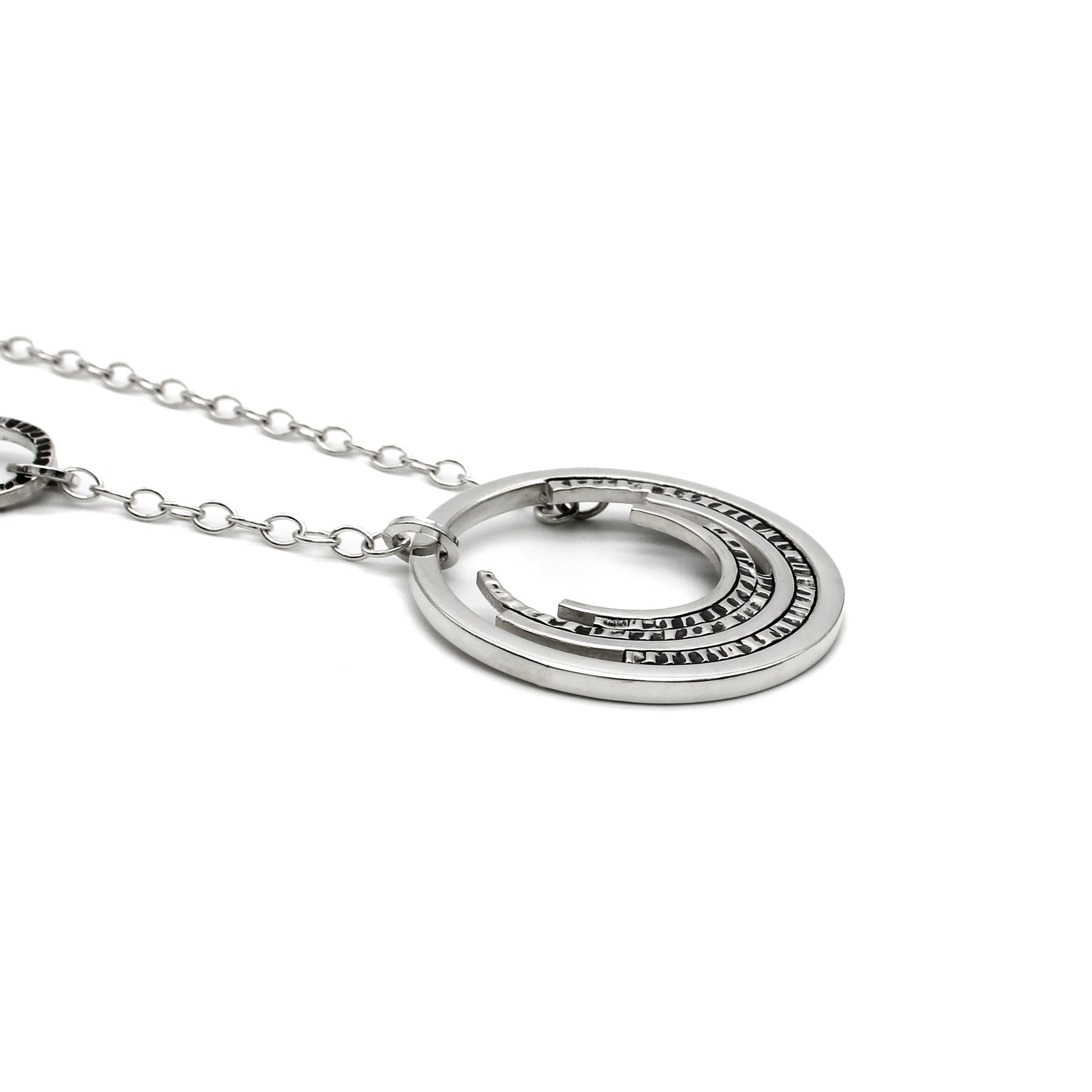 Contemporary sterling silver necklace with a prominent outer circle surrounded by five descending, staggered inner circles. The inner circles showcase varying textures, some chisel-textured and blackened for enhanced visual contrast. Suspended from a sterling silver cable chain, the pendant is complemented by four irregularly sized and textured rings positioned 1.5 inches up the chain on the right side. Necklace is laying flat, and view is from the side. 