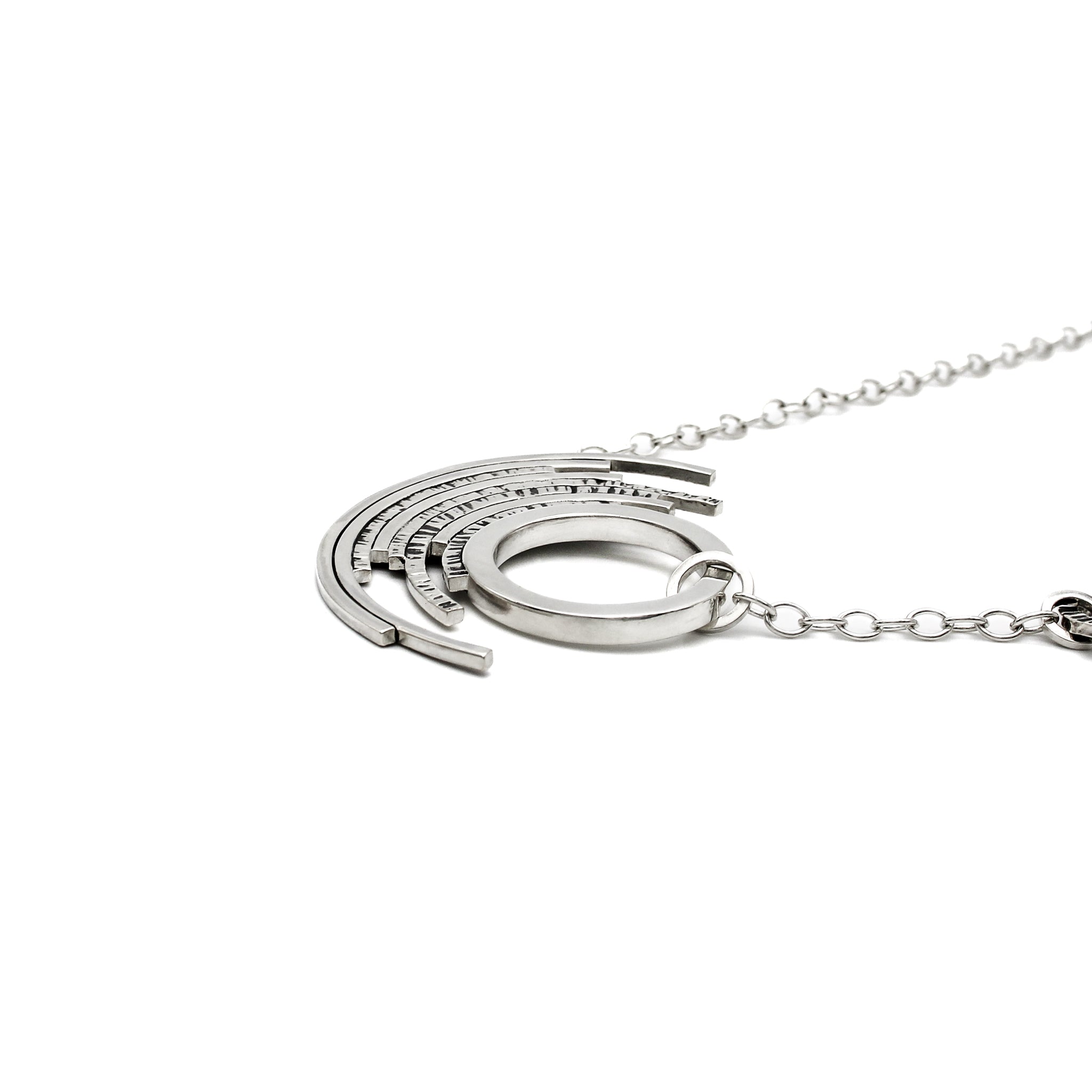 A handmade sterling silver asymmetrical pendant with a single thick circle surrounded by 7 outer concentric partial circles, some with featuring a chisel texture. Necklace is laying flat, view is across the front of piece from the right side. 