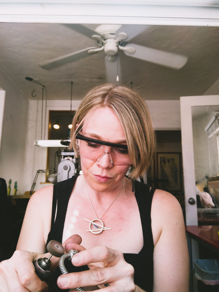 A front view of a woman seated at a jewelry bench. She wears a black apron and safety goggles, holding a silver bracelet in her left hand. She is engaged in the polishing process, operating a flexshaft in her right hand. In the background, a well-equipped jewelry studio can be seen. 